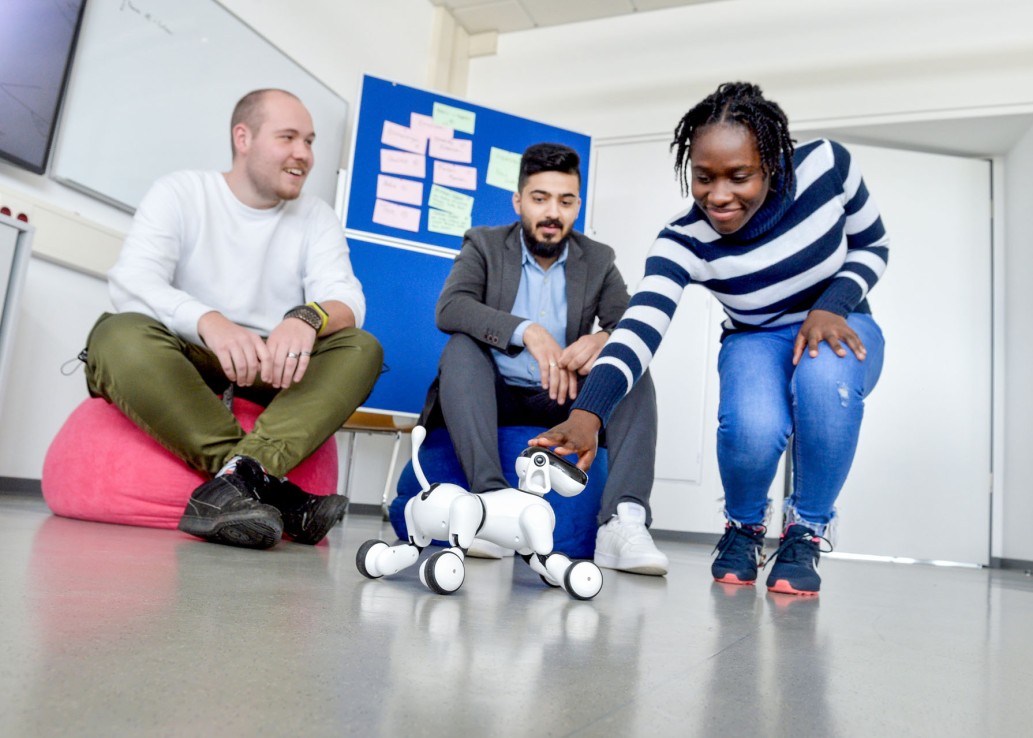 Zwei junge Männer sitzen in einem Raum auf Sitzsäcken. Eine junge Frau beugt sich lächelnd vor und streichelt einem Roboterhund über den Kopf.