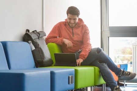 Studierender sitzt auf dem Flur der Hochschule und schaut auf ein Tablet.