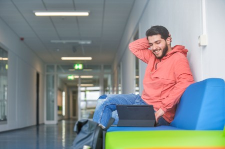 Studierender sitzt auf dem Flur der Hochschule und schaut auf ein Tablet.