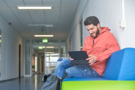 Studierender sitzt auf dem Flur der Hochschule und schaut auf ein Tablet.