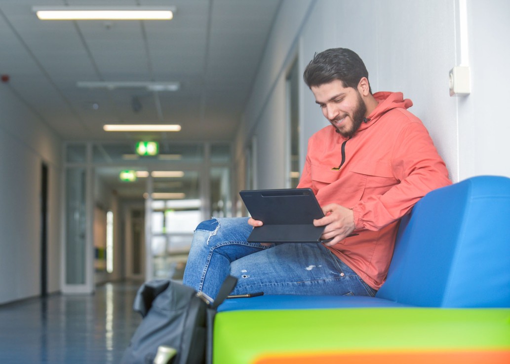 Studierender sitzt auf dem Flur der Hochschule und schaut auf ein Tablet.