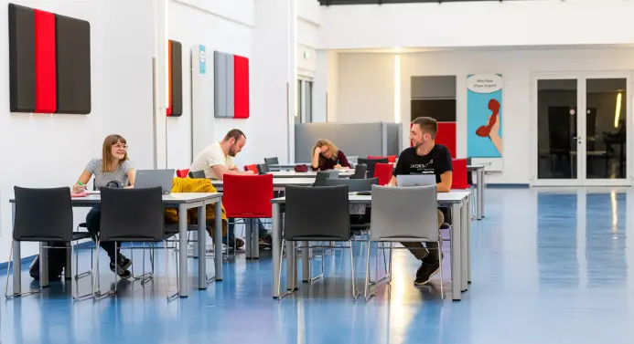 Studierende sitzen in einem großen offenen Raum an Tischen und machen sich Notizen.