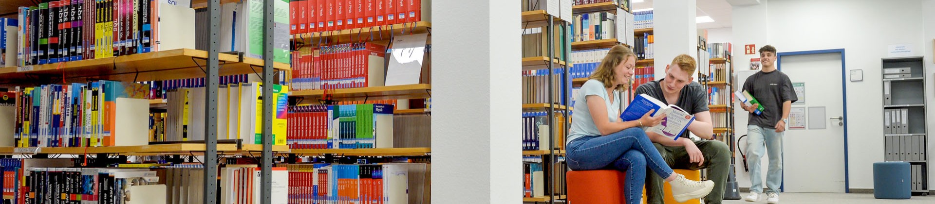 Zwei Personen sitzen in einer Bibliothek und schauen zusammen in ein Buch. Eine dritte Person läuft im Hintergrund mit zwei Büchern in der Hand vorbei.