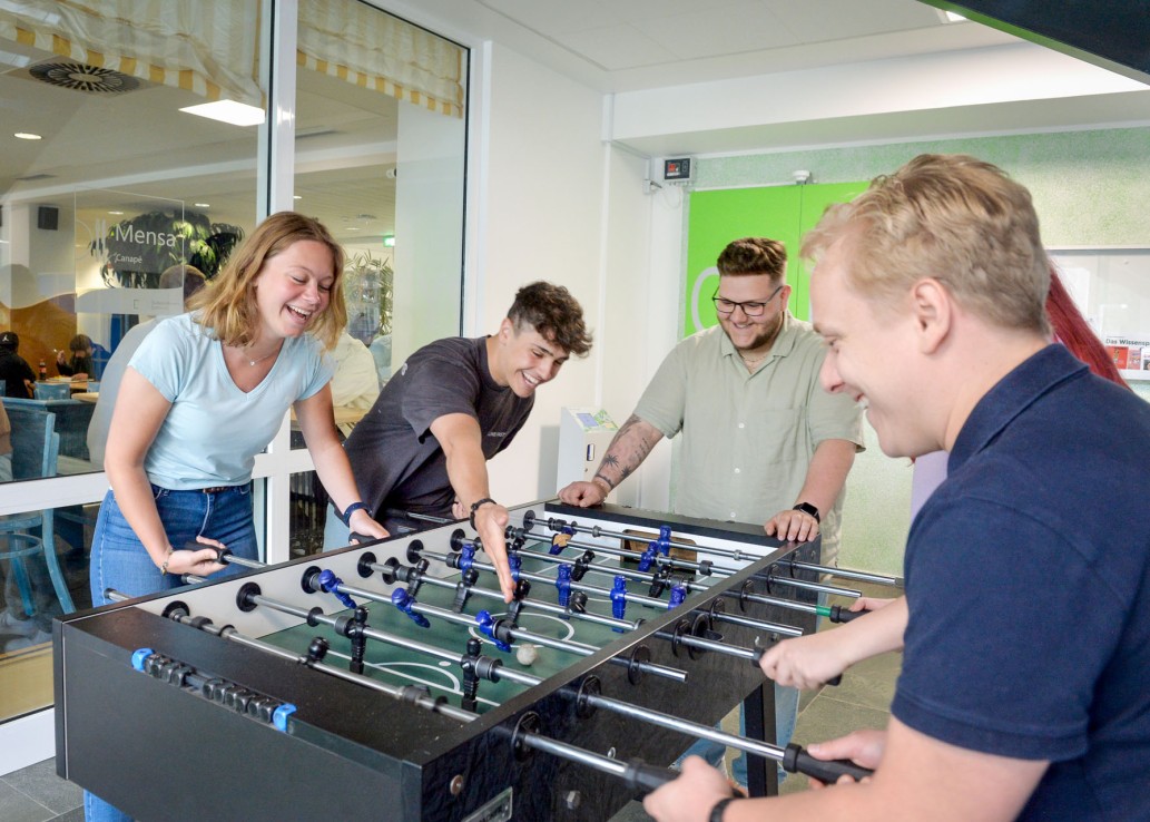 Gruppe junger Frauen und Männer spielen gemeinsam an einem Kickertisch und lachen.