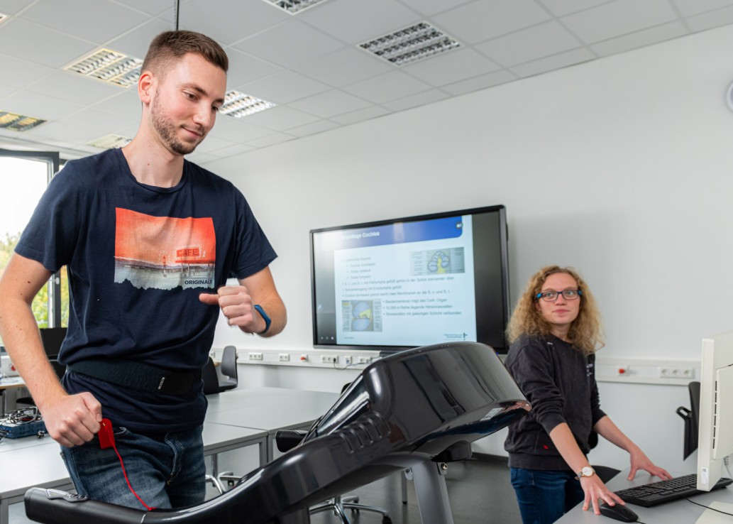 Ein junger Mann läuft auf einem Laufband. Er trägt einen Messgürtel und Kabel um seine Hüfte. Eine junge Frau steht an einem PC daneben und schaut in Richtung des Mannes.