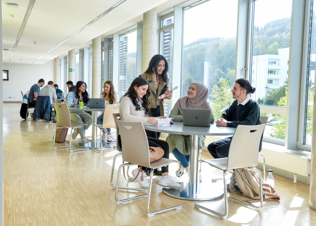 In einem hellen Flur vor einer großen Fensterfront sitzen junge Frauen und Männer in Gruppen an drei hintereinander stehenden Tischen, arbeiten an Laptops und unterhalten sich.