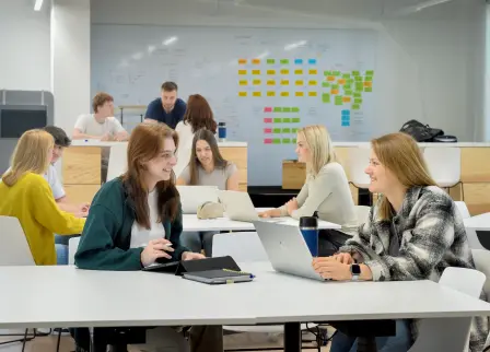 Zwei Frauen sitzen sich an einem Tisch im dpm-Workspace der FH Südwestfalen lächelnd gegenüber, auf dem Tisch vor ihnen liegen ein Tablet und einen Laptop. Im Hintergrund sind weitere Gruppen an Tischen und viele bunte an der Wand angebrachte Post-its zu sehen.