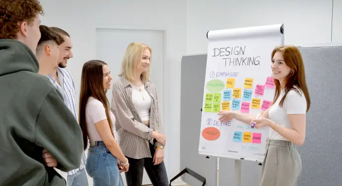 Eine Frau steht vor einer Gruppe Frauen und Männer und zeigt erklärend auf ein Post-It, das auf einer Flipchart unter der Überschrift