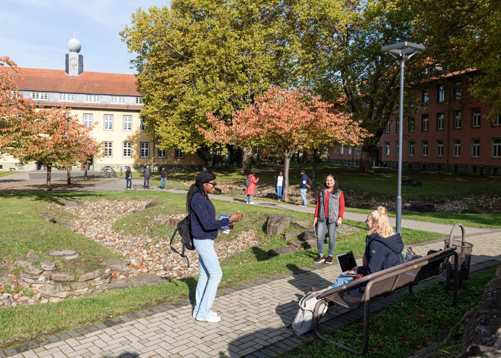 Drei Gruppen junger Menschen stehen draußen auf gepflasterten Wegen zwischen zwei Wiesen zusammen und unterhalten sich. Um sie herum sind viele Bäume mit grün-rotem Laub und zwei Gebäude - eines davon mit einem kleinen Türmchen mit Uhr auf dem Dach.