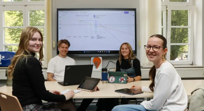 Vier Studierende sitzen an einem Tisch und lächeln in die Kamera. Vor ihnen auf dem Tisch liegen ein Laptop, Tablets, ein Block und Stift. Im Hintergrund wird an einem Bildschirm eine physikalische Zeichnung angezeigt.