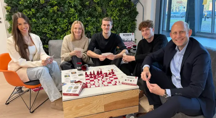 Studierende und Professor sitzen an einem Tisch, auf dem ein Memory und ein Schachbrett liegen, und halten Spielkarten in der Hand.