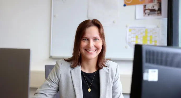 Eine Frau sitzt an einem Schreibtisch vor einem Computer und lächelt in die Kamera.