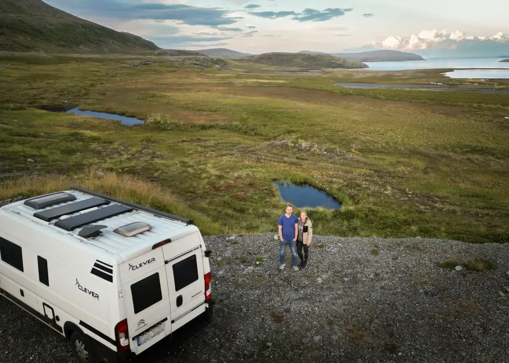 Ein Mann und eine Frau stehen neben einem Camper umgeben von einer schönen Landschaft.