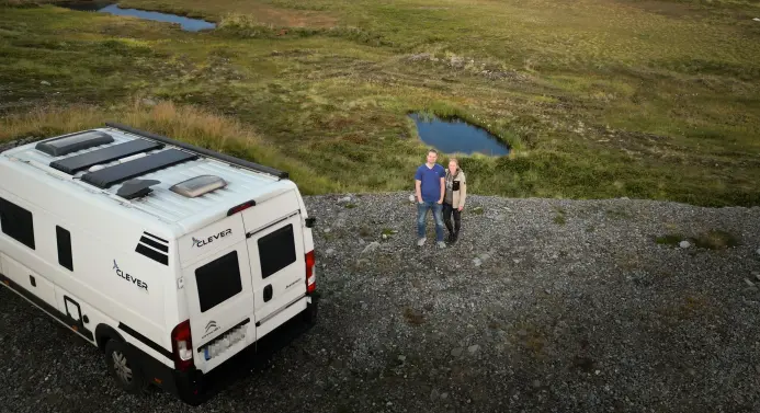 Ein Mann und eine Frau stehen neben einem Camper umgeben von einer schönen Landschaft.