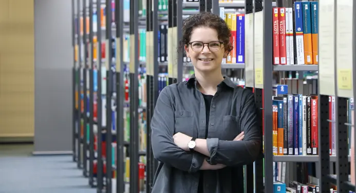 Prof. Dr. Kristina Kusanke lehnt an einem Bücherregal in einer Bibliothek.