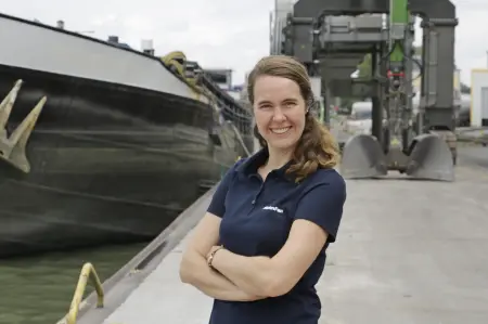 Eine Frau steht an einem Hafen, links ist ein Schiff zu sehen, im Hintergrund eine Krananlage.