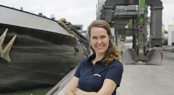 Eine Frau steht an einem Hafen, links ist ein Schiff zu sehen, im Hintergrund eine Krananlage.