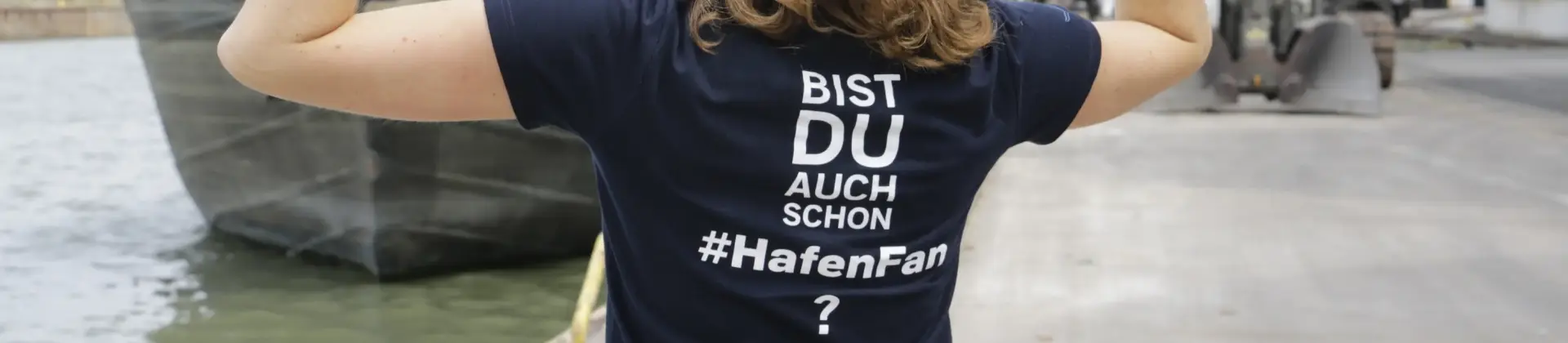 Eine Frau steht an einem Hafen mit dem Rücken zur Kamera gedreht. Auf ihrem T-Shirt steht: 