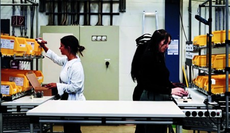 Zwei Frauen stehen in einem mit Tischen geformten U und bauen Produkte zusammen. An der rechten und linken Seite sind jeweils mehrere Boxen aufgereiht.